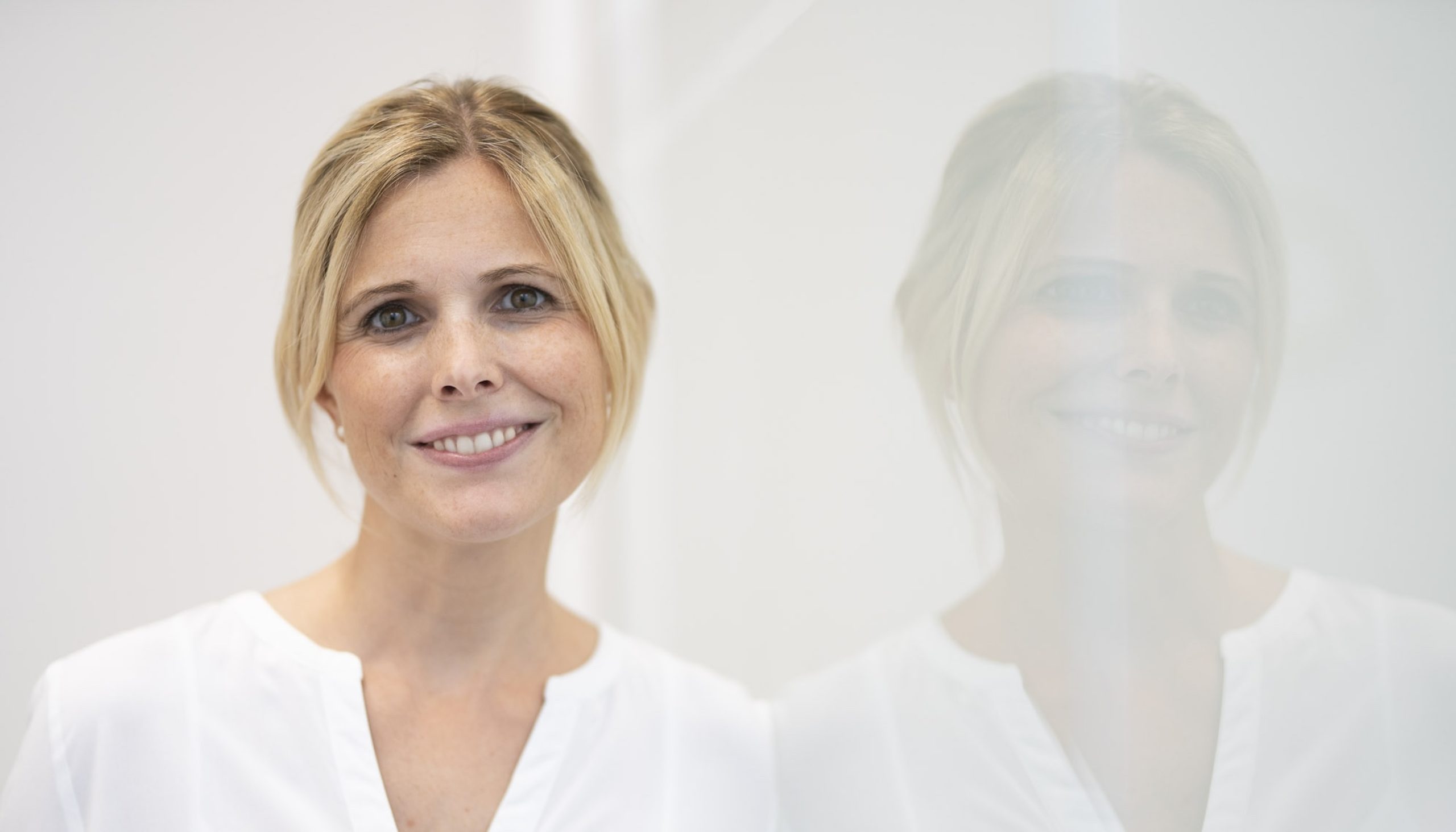 Freundlich lächelnde Frau mit hellblondem Haar und weißer Bluse, stehend neben einer Glaswand.