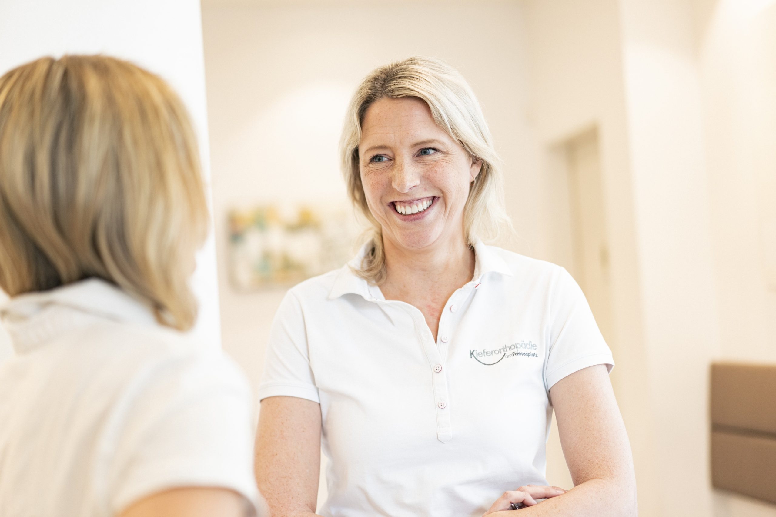 Zwei Mitarbeiterinnen der Kieferorthopädie am Friesenplatz unterhalten sich freundlich; eine lächelnde Frau in weißem Polohemd mit Praxislogo steht im Vordergrund.