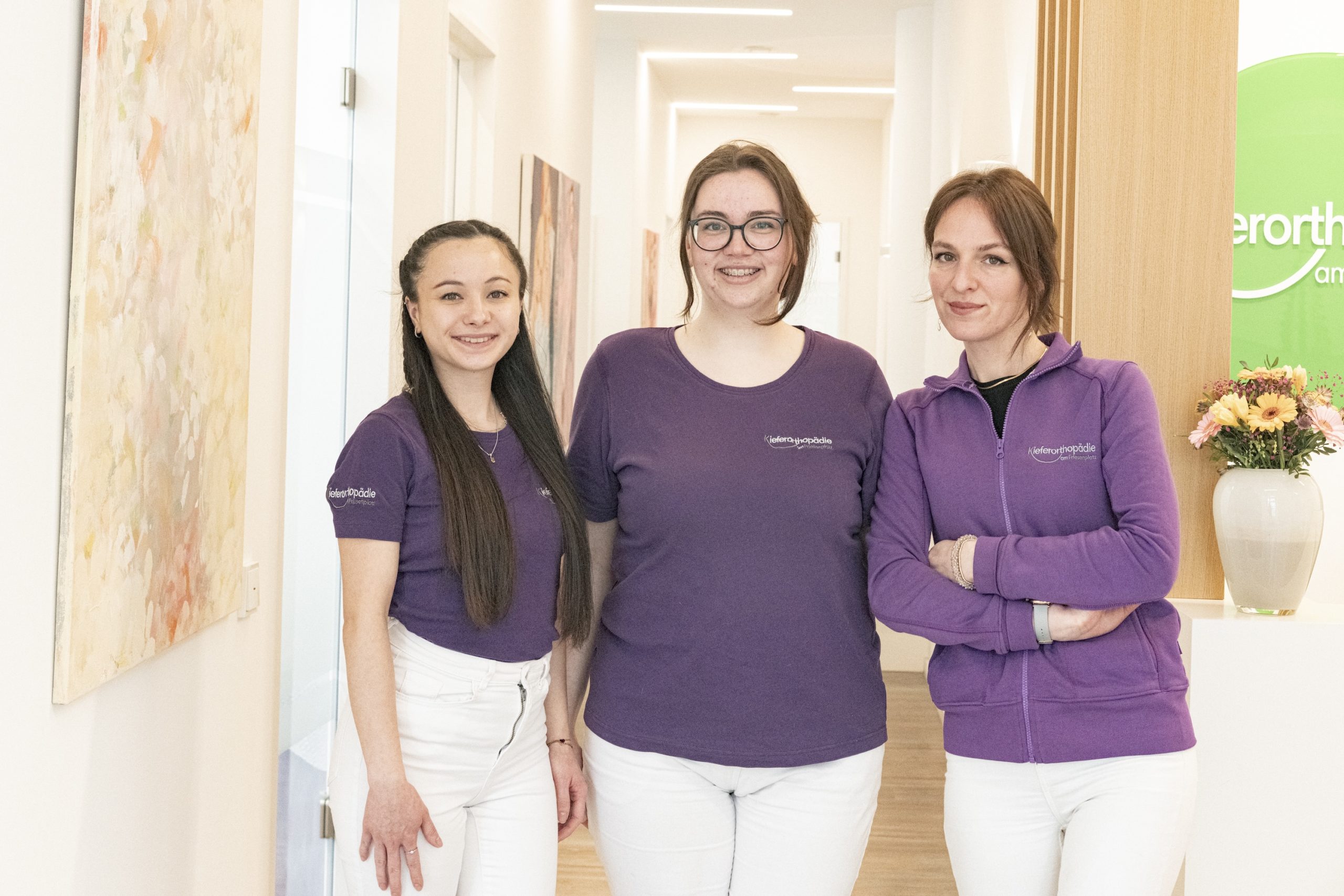 Drei Mitarbeiterinnen der Kieferorthopädie am Friesenplatz stehen im Flur der Praxis, tragen lilafarbene Oberteile mit Praxislogo und lächeln in die Kamera.