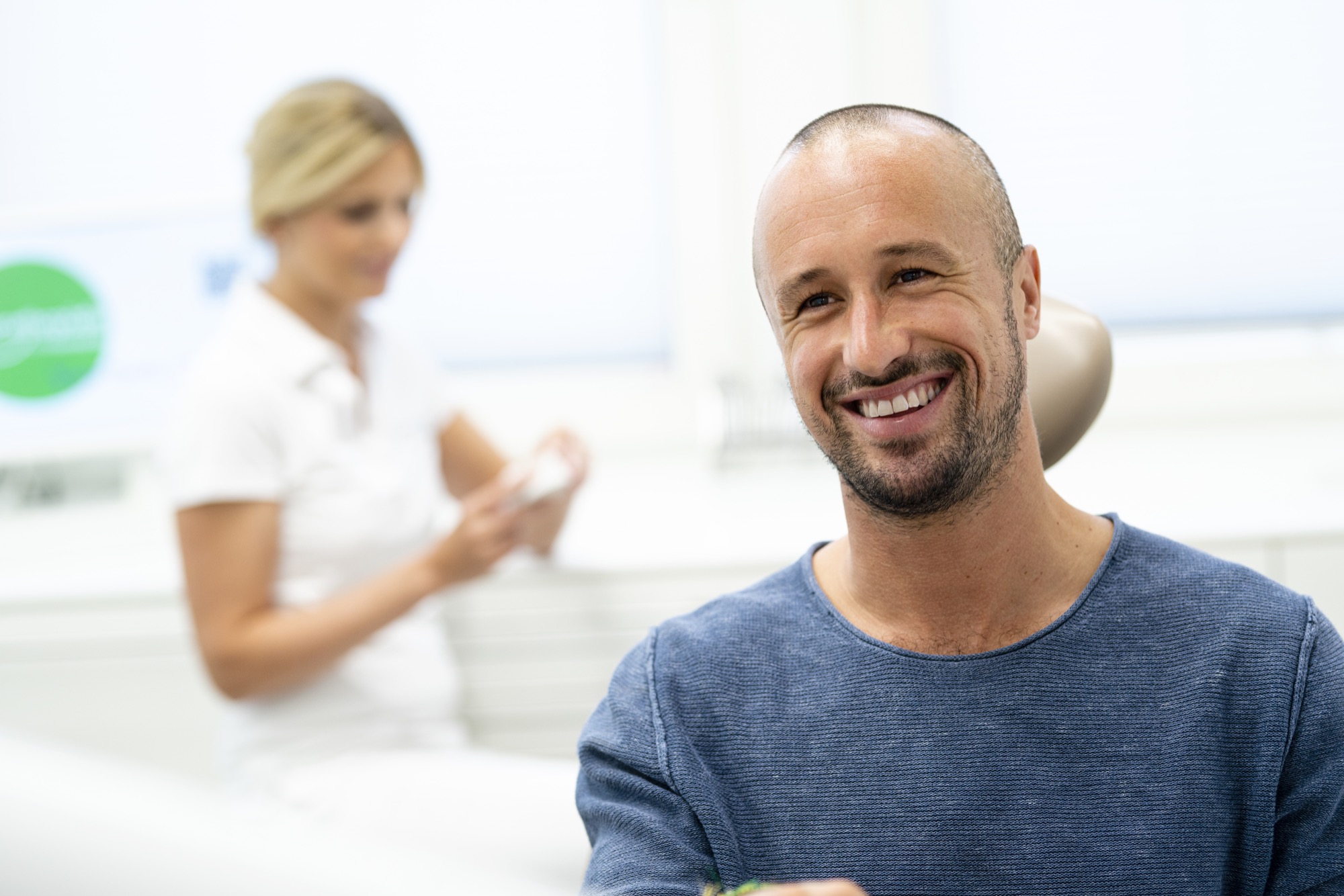 Lächelnder Patient sitzt im Behandlungsstuhl, während die Kieferorthopädin im Hintergrund ein Zahnmodell vorbereitet.