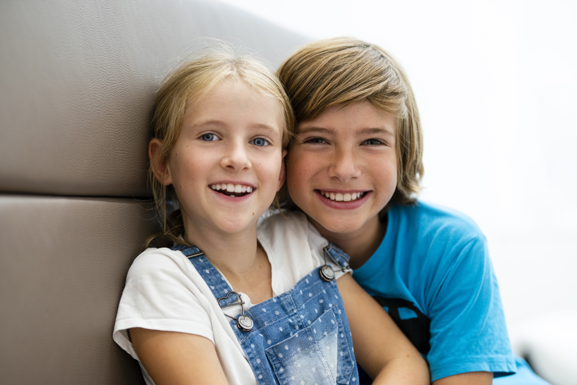 Zwei fröhliche Kinder sitzen nebeneinander und lächeln in die Kamera, im Hintergrund eine helle Praxisumgebung.