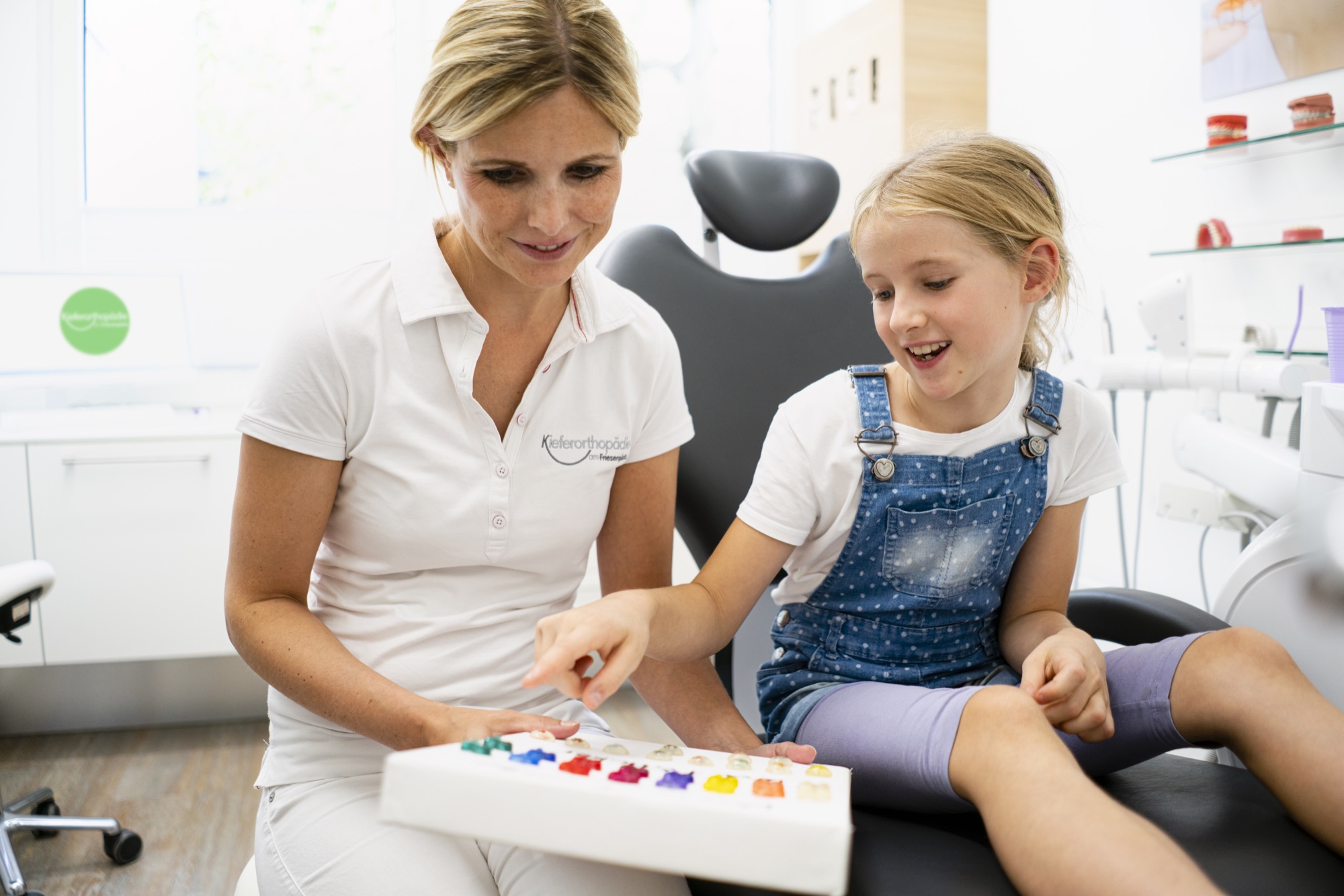 Kieferorthopädin zeigt einem Mädchen verschiedene bunte Zahnspangen, das Kind sitzt im Behandlungsstuhl und wählt interessiert eine Farbe aus.