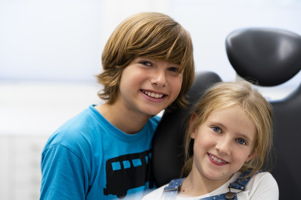 Zwei Kinder sitzen nebeneinander im Behandlungsstuhl der Kieferorthopädie und lächeln in die Kamera.