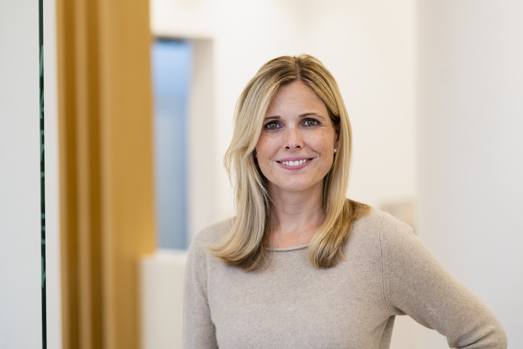 Freundlich lächelnde Frau mit hellblondem Haar in beigefarbenem Pullover, stehend in moderner Praxisumgebung.